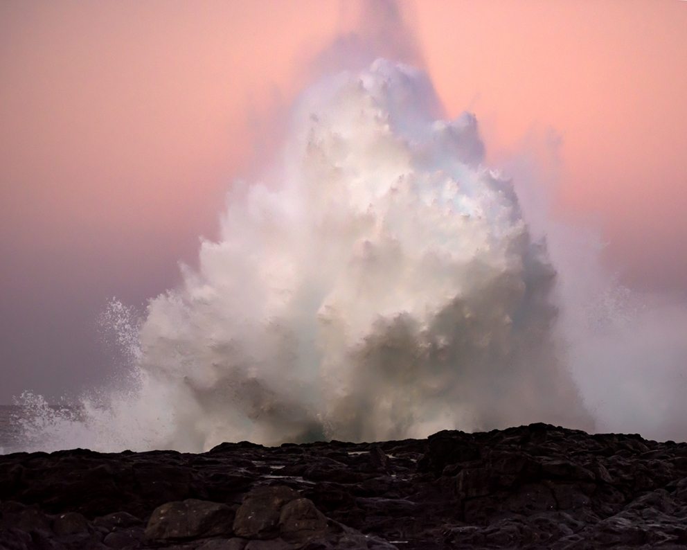 A photograph of a giant wave crashing against the darkened shore just after sunset with a pink sky.